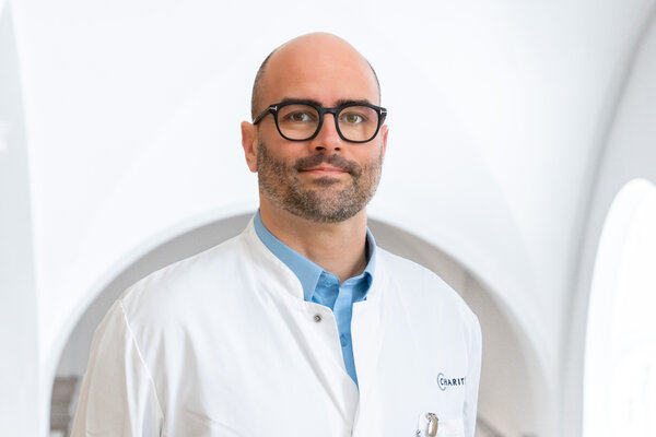 Ein Arzt mit Brille steht in einem hellen, modernen Raum. Er trägt einen weißen Kittel über einem blauen Hemd. Der Hintergrund wirkt freundlich und einladend, mit großen Fenstern und sanften Lichtverhältnissen.