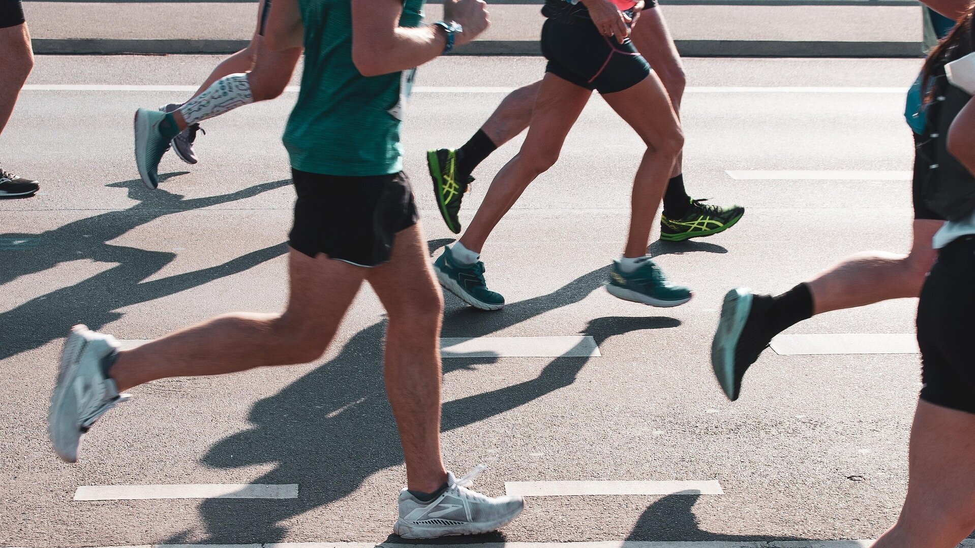 Eine Gruppe von Läufern mit kurzen Hosen und Sportschuhen läuft auf einer Straße. Die Beine sind in Bewegung, und die Schatten der Läufer fallen auf den Asphalt. Die Szene vermittelt Dynamik und sportliche Aktivität.