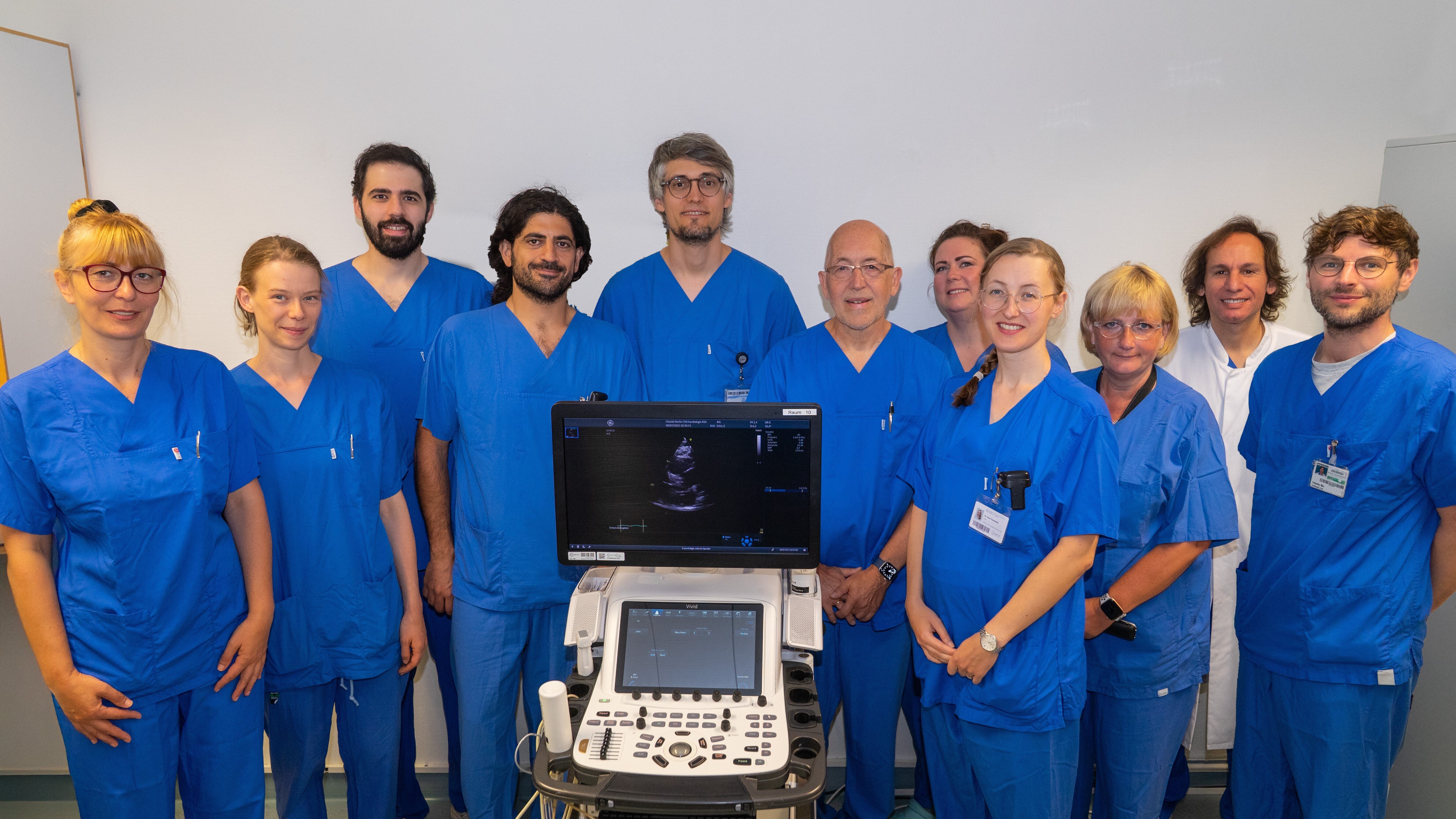 Eine Gruppe von zehn medizinischen Fachkräften in blauen Krankenhauskitteln steht vor einem Ultraschallgerät. Die Personen lächeln, während sie für das Foto posieren. Im Hintergrund sind neutrale Wände, die den Fokus auf die Gruppe lenken.