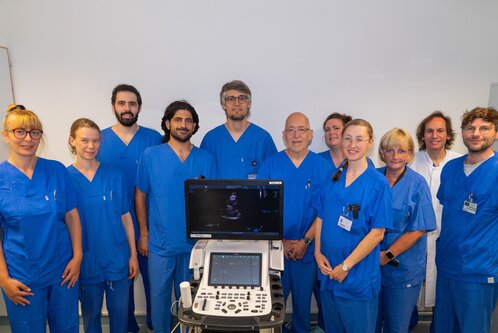 Eine Gruppe von zehn medizinischen Fachkräften in blauen Krankenhauskitteln steht vor einem Ultraschallgerät. Die Personen lächeln, während sie für das Foto posieren. Im Hintergrund sind neutrale Wände, die den Fokus auf die Gruppe lenken.