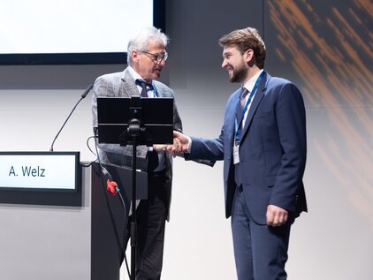 Zwei Männer schütteln sich die Hände auf einer Bühne. Einer trägt eine Brille und eine graue Jacke, der andere einen dunklen Anzug. Vor ihnen steht ein Rednerpult mit dem Namen "A. Welz". Im Hintergrund ist ein helles, neutrales Display sichtbar.