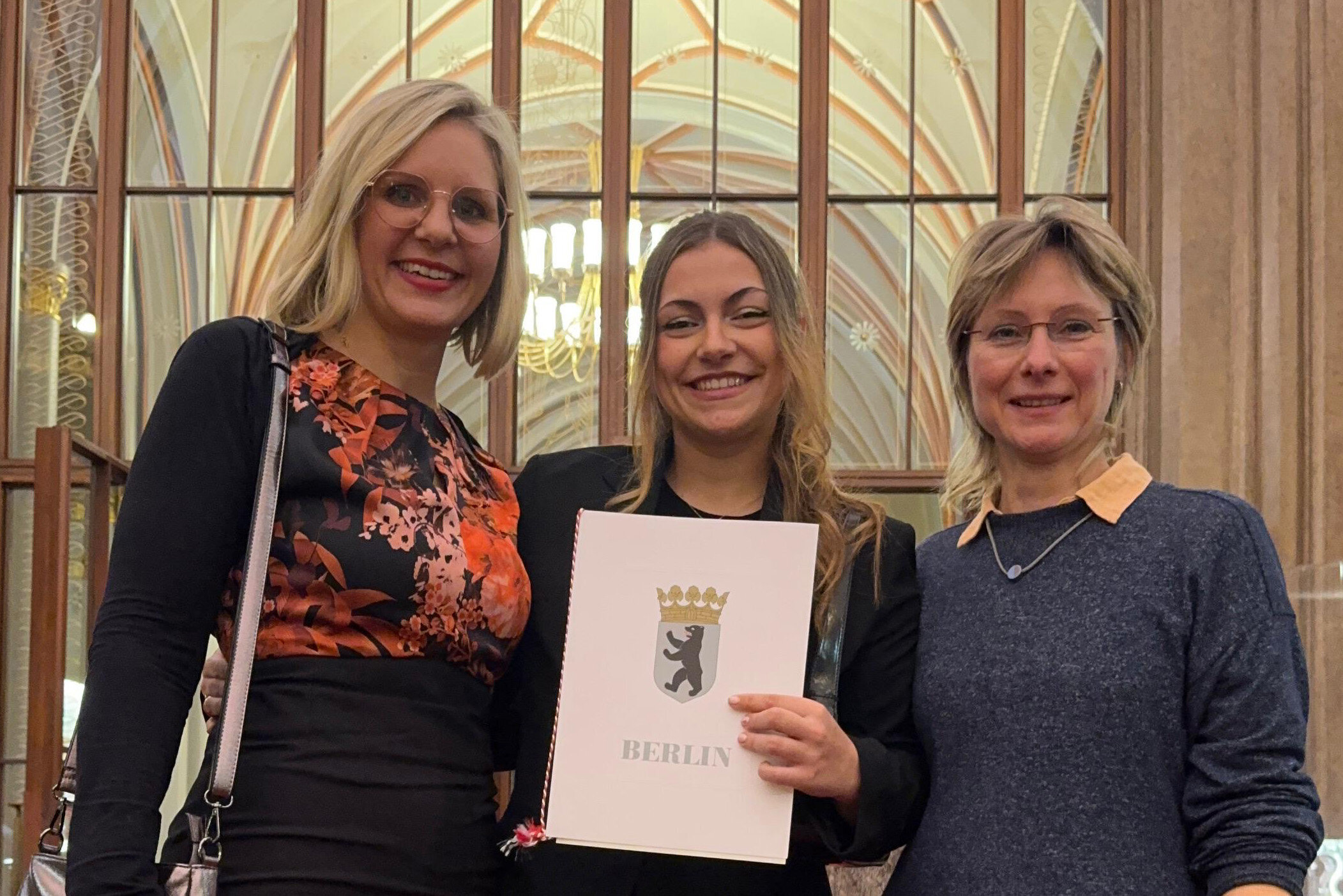 Drei Frauen stehen lächelnd vor einer eleganten Kulisse. Die mittlere Frau hält ein Dokument mit dem Aufdruck „BERLIN“ und einem Wappen. Alle tragen ansprechende Kleidung und wirken stolz zu sein. Im Hintergrund sind Fenster und lichtdurchflutete Details sichtbar.