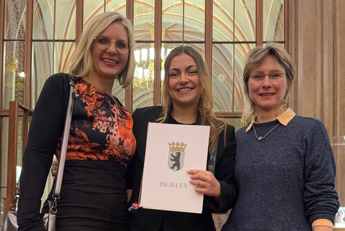 Drei Frauen stehen lächelnd vor einer eleganten Kulisse. Die mittlere Frau hält ein Dokument mit dem Aufdruck „BERLIN“ und einem Wappen. Alle tragen ansprechende Kleidung und wirken stolz zu sein. Im Hintergrund sind Fenster und lichtdurchflutete Details sichtbar.