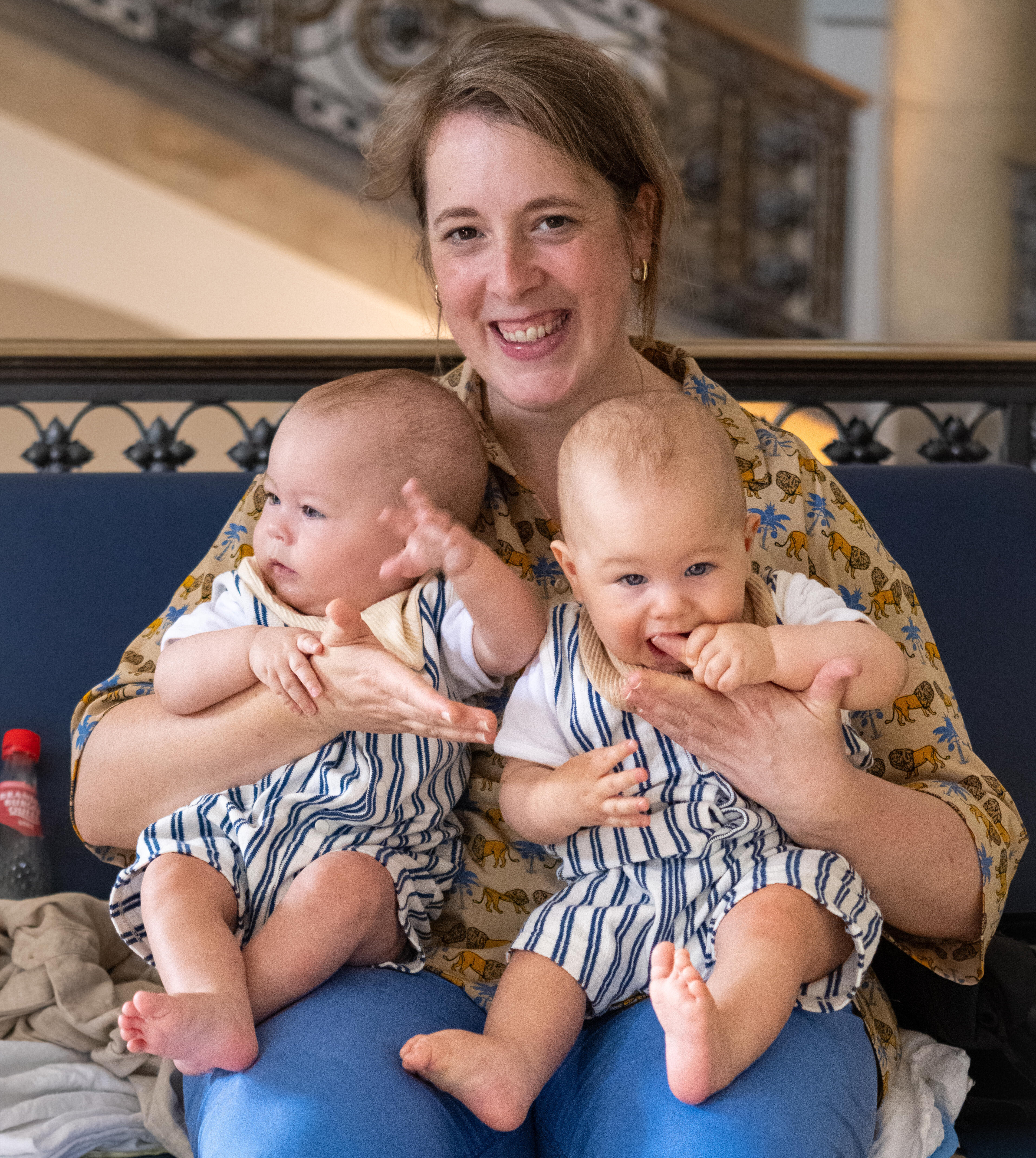 Eine lächelnde Frau sitzt auf einer Bank und hält zwei Säuglinge auf ihren Knien. Die Babys tragen gestreifte Overalls und scheinen glücklich zu sein. Die Umgebung ist hell und freundlich.