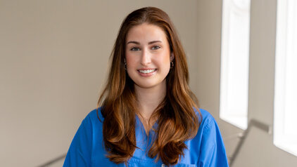 Eine junge Frau mit langen, braunen Haaren lächelt freundlich in die Kamera. Sie trägt ein blaues medizinisches Outfit und steht in einem hellen Raum mit großen Fenstern im Hintergrund.