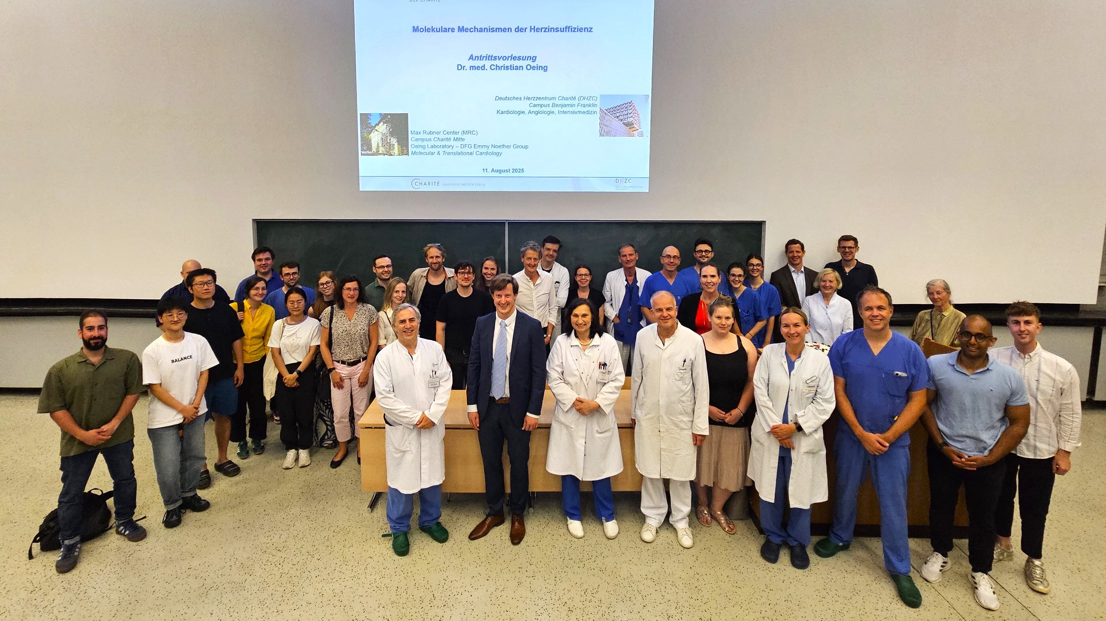 Gruppenfoto von Teilnehmern einer medizinischen Veranstaltung zum Thema "Molekulare Mechanismen der Herzinsuffizienz". Im Vordergrund stehen Ärzte in weißen Kitteln und Anzug, während im Hintergrund eine gemischte Gruppe von Fachleuten und Studierenden versammelt ist. An der Wand ist eine Präsentation sichtbar.