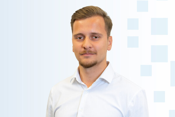 Ein junger Mann mit kurzen Haaren und einem leichten Bart trägt ein helles Hemd. Er steht vor einem hellblauen Hintergrund mit geometrischen Formen. Der Gesichtsausdruck ist neutral und selbstbewusst.