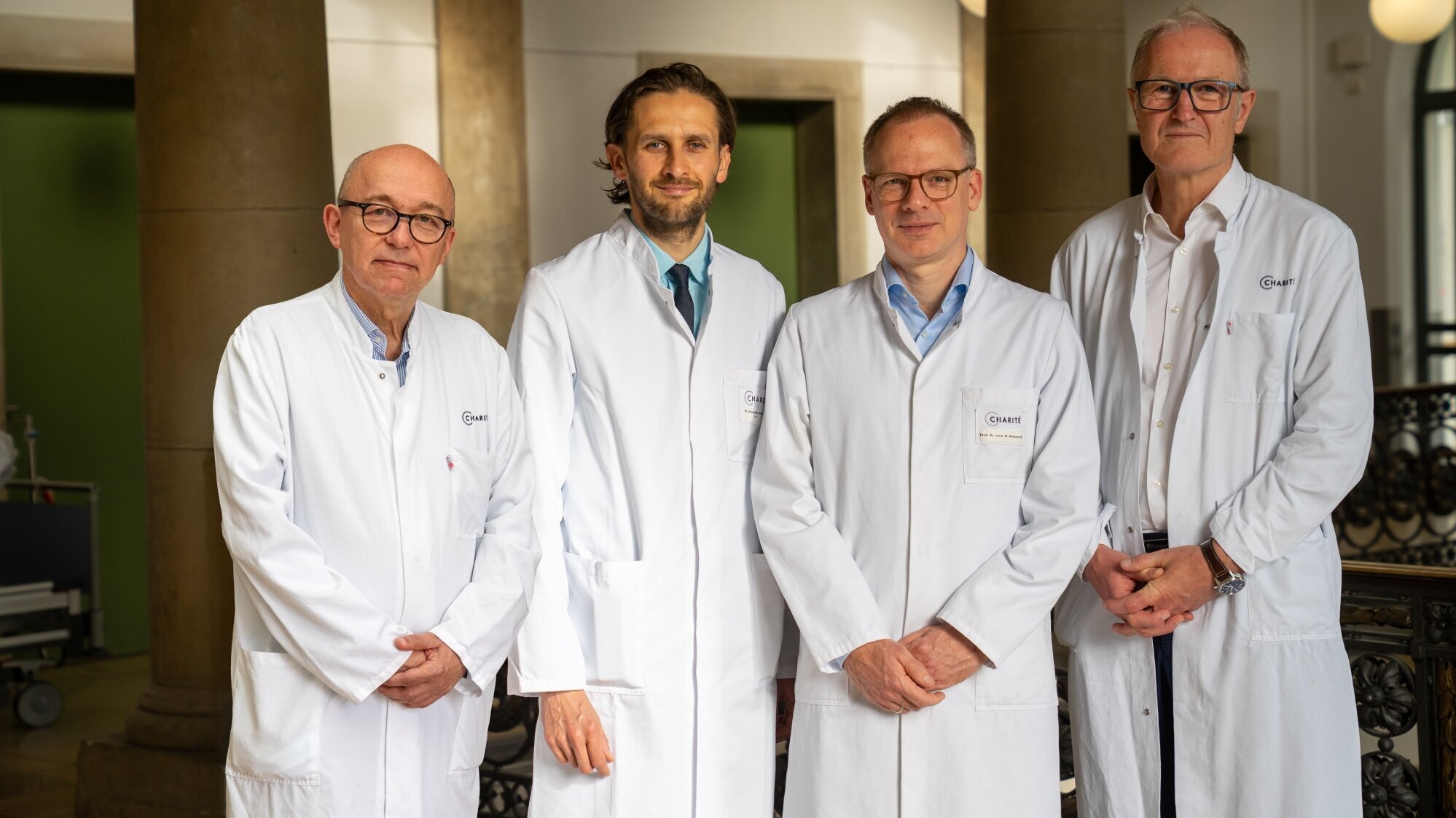 Vier Ärzte in weißen Kitteln stehen nebeneinander in einem Gebäude. Sie blicken freundlich in die Kamera. Der Hintergrund ist hell mit historischen Elementen. Die Ärzte repräsentieren eine medizinische Einrichtung oder eine Konferenz.