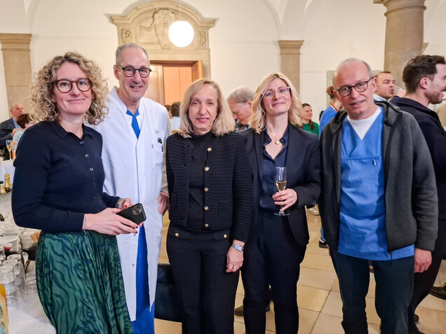Fünf Personen stehen in einem hellen Raum, umgeben von Feierlichkeiten. Zwei Frauen tragen elegante Kleidung, eine hält ein Glas. Ein Arzt in weißem Kittel steht in der Mitte. Weitere Gäste sind im Hintergrund sichtbar, was auf eine gesellige Veranstaltung hinweist.