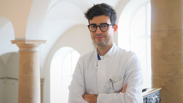 Ein junger Arzt steht in einem hellen Raum mit großen Fenstern. Er trägt einen weißen Kittel und hat die Arme vor der Brust verschränkt. Sein Gesichtsausdruck ist freundlich und selbstbewusst. Im Hintergrund sind Säulen und ein elegantes Geländer zu sehen.