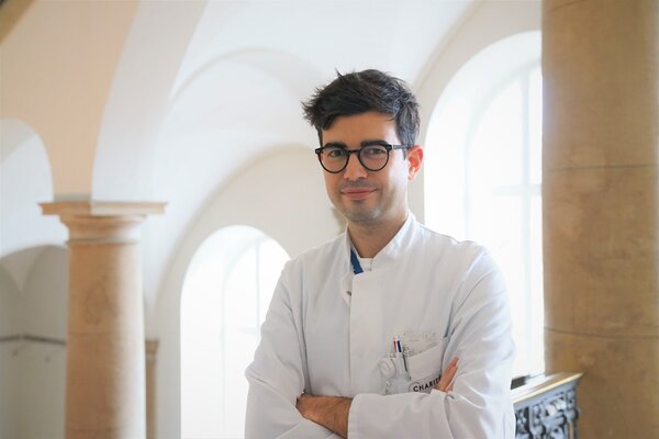 Ein junger Arzt steht in einem hellen Raum mit großen Fenstern. Er trägt einen weißen Kittel und hat die Arme vor der Brust verschränkt. Sein Gesichtsausdruck ist freundlich und selbstbewusst. Im Hintergrund sind Säulen und ein elegantes Geländer zu sehen.