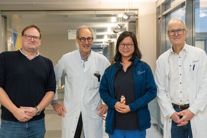 Vier Personen stehen in einem Krankenhausflur. Eine Person trägt einen weißen Kittel, zwei weitere Personen sind in blauen Jacken gekleidet, und eine Person trägt ein schwarzes Oberteil. Alle lächeln und posieren freundlich. Im Hintergrund sind Gänge und Deckenleuchten sichtbar.