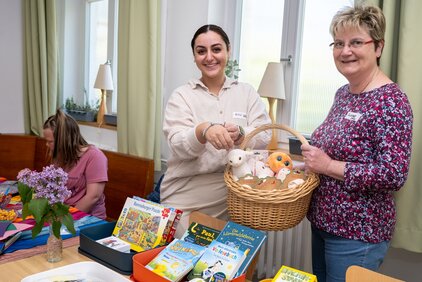 Eine junge Frau und eine ältere Frau lächeln, während sie einen Korb mit Spielzeug und Stofftieren präsentieren. Auf dem Tisch sind verschiedene Bücher und ein Blumenstrauß zu sehen. Im Hintergrund sitzt eine andere Frau, die an einem Tisch beschäftigt ist.