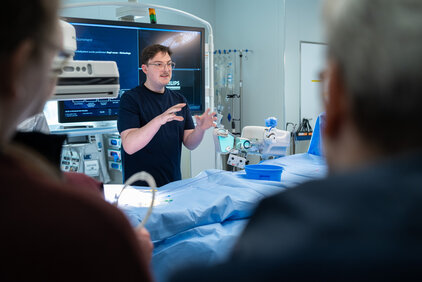 Ein junger Mann erklärt anschaulich ein medizinisches Thema in einem modernen Operationssaal. Auf einem Bildschirm im Hintergrund sind Informationen sichtbar. Vor ihm stehen zwei Zuhörer, die aufmerksam zuhören und Notizen machen. Der Tisch ist mit einer blauen Abdeckung ausgestattet.