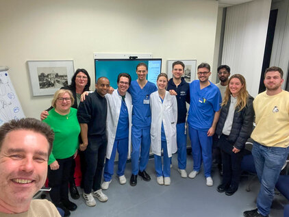 Eine Gruppe von zwölf Personen steht in einem Raum. Sie tragen überwiegend medizinische Arbeitskleidung, darunter blaue und weiße Kittel. Lächelnde Gesichter deuten auf eine freundliche Atmosphäre hin. Im Vordergrund ist eine Person zu sehen, die in die Kamera lächelt.