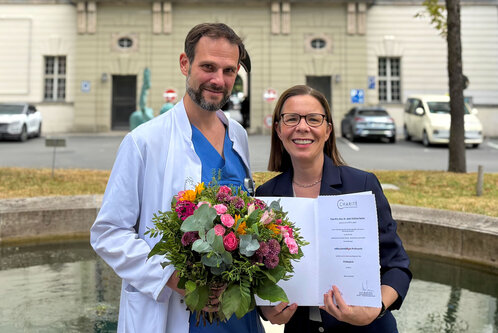 Ein Mann in einem weißen Kittel hält einen Blumenstrauß, während eine Frau ihm stolz ein Dokument präsentiert. Beide lächeln in die Kamera. Im Hintergrund ist eine Gebäudefassade und Autos sichtbar, die den feierlichen Anlass unterstreichen.