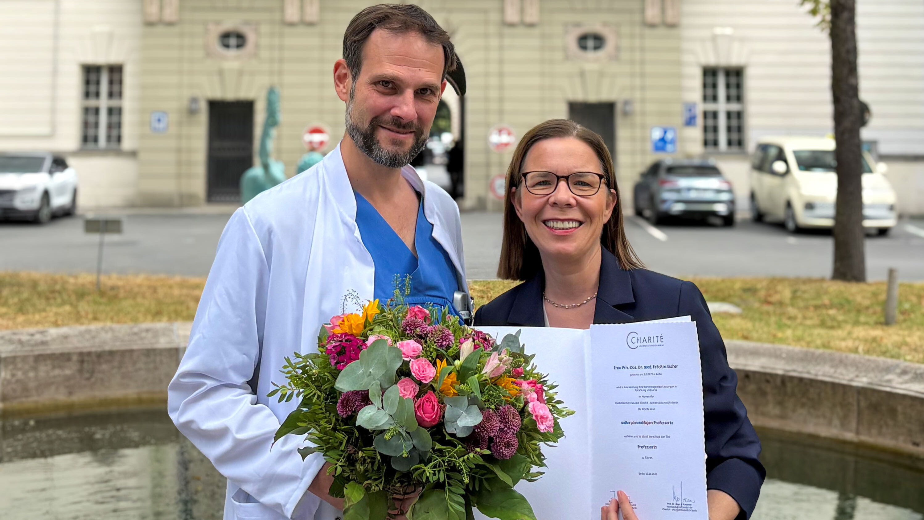 Ein Mann in einem weißen Kittel hält einen Blumenstrauß, während eine Frau ihm stolz ein Dokument präsentiert. Beide lächeln in die Kamera. Im Hintergrund ist eine Gebäudefassade und Autos sichtbar, die den feierlichen Anlass unterstreichen.