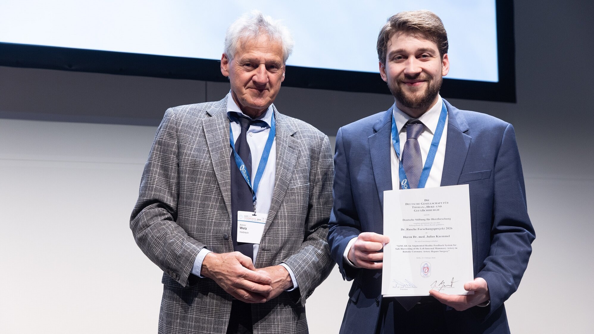 Zwei Männer stehen nebeneinander auf einer Bühne. Der ältere Mann, in einem karierten Anzug, lächelt und hält eine Urkunde. Der jüngere Mann, gekleidet in einen Anzug mit einer Krawatte, zeigt stolz die Urkunde, die er gerade erhalten hat.