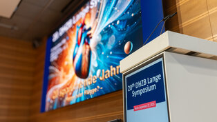 Ein Rednerpult steht vor einer Leinwand, die für das 20. DHZB Lange Symposium am 27. September 2025 in Berlin beworben wird. Im Hintergrund ist ein buntes Bild eines Herzens und planetarischer Motive zu sehen.