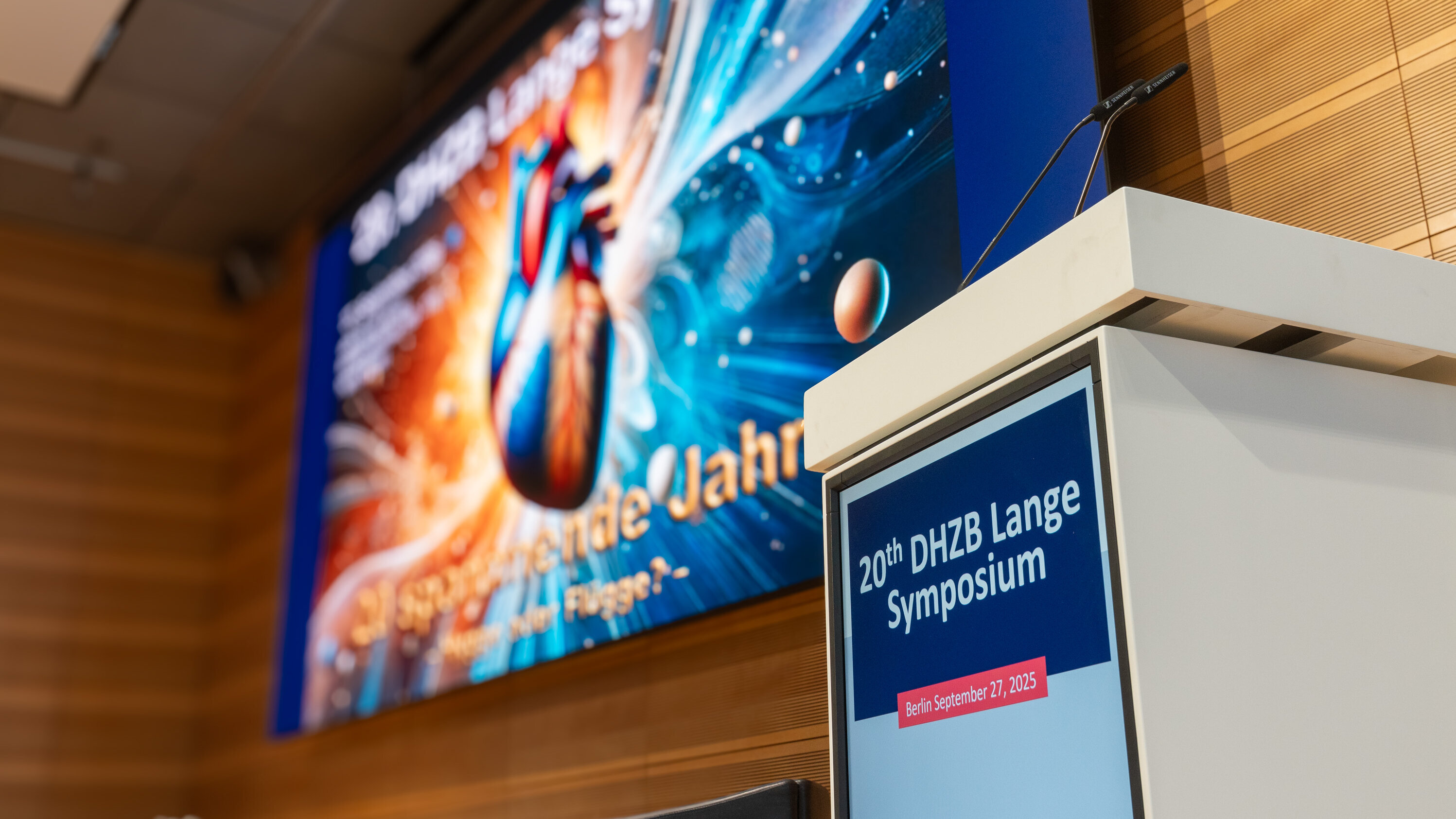 Ein Rednerpult steht vor einer Leinwand, die für das 20. DHZB Lange Symposium am 27. September 2025 in Berlin beworben wird. Im Hintergrund ist ein buntes Bild eines Herzens und planetarischer Motive zu sehen.