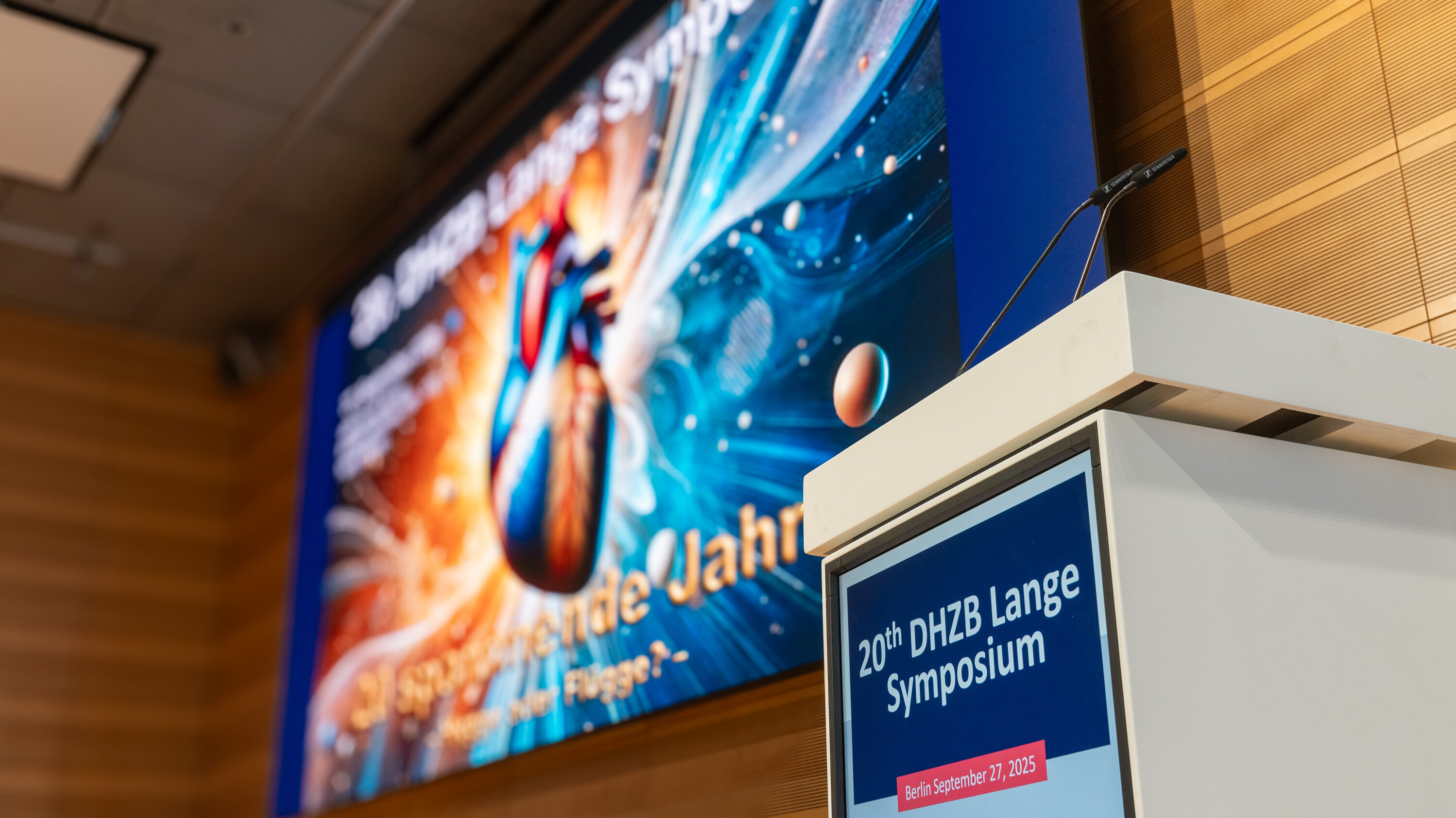 Ein Rednerpult steht vor einer Leinwand, die für das 20. DHZB Lange Symposium am 27. September 2025 in Berlin beworben wird. Im Hintergrund ist ein buntes Bild eines Herzens und planetarischer Motive zu sehen.