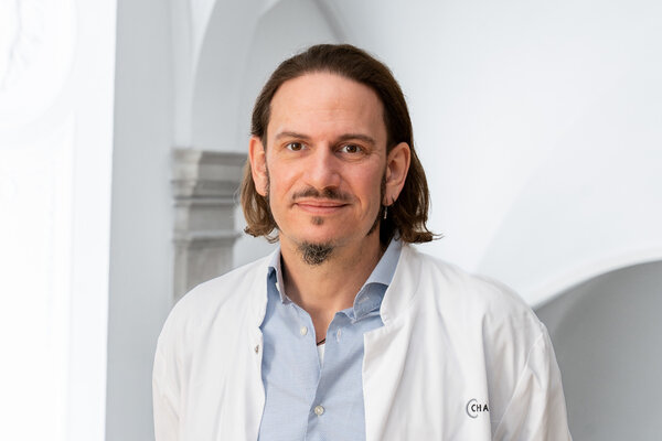 Ein männlicher Arzt steht lächelnd in einer hellen Umgebung. Er trägt einen weißen Kittel über einem hellblauen Hemd und hat lange, dunkle Haare. Der Hintergrund ist unscharf, bietet jedoch einen Eindruck von Professionalität und Freundlichkeit.