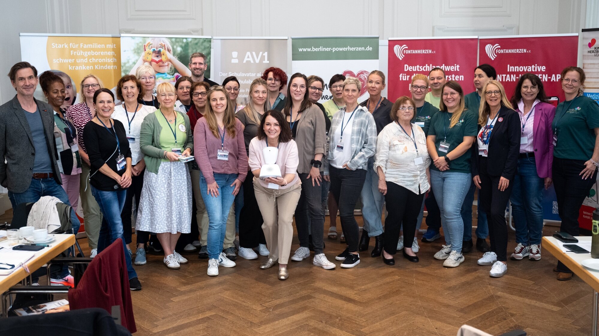 Eine Gruppe von etwa 30 Personen steht in einem Raum, lächelt und posiert für ein Gruppenfoto. Im Hintergrund sind Werbebanner sichtbar. Die Personen tragen unterschiedliche Kleidung und scheinen an einer Veranstaltung teilzunehmen, die gemeinschaftlich und freundlich wirkt.