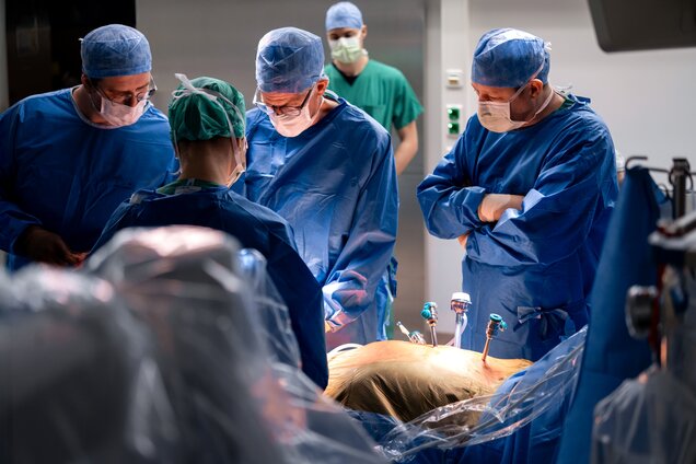 Eine Gruppe von Chirurgen in blauer OP-Kleidung und Masken konzentriert sich auf einen chirurgischen Eingriff. Sie stehen um einen Patienten, der auf einem Operationstisch liegt und mit Instrumenten verbunden ist. Die Atmosphäre ist ernst und professionell.