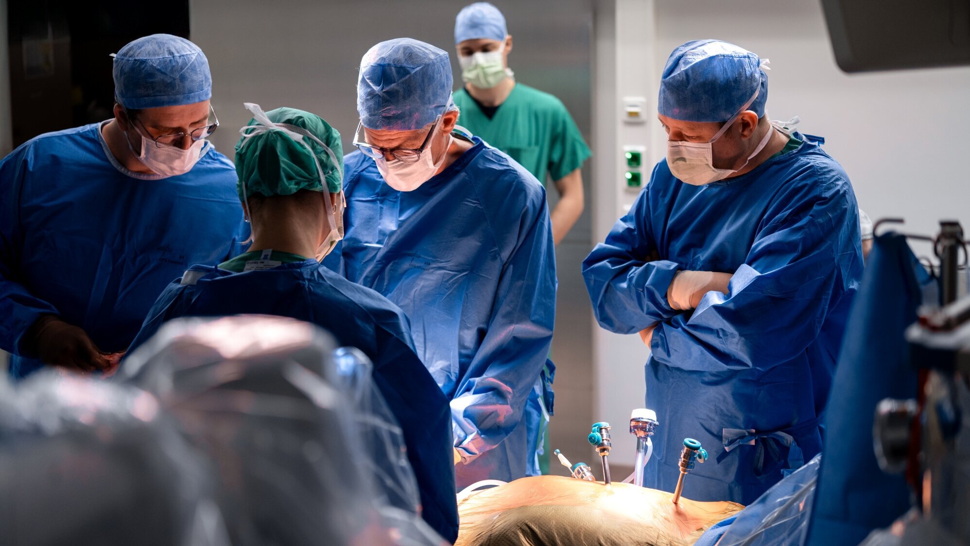 Eine Gruppe von Chirurgen in blauer OP-Kleidung und Masken konzentriert sich auf einen chirurgischen Eingriff. Sie stehen um einen Patienten, der auf einem Operationstisch liegt und mit Instrumenten verbunden ist. Die Atmosphäre ist ernst und professionell.