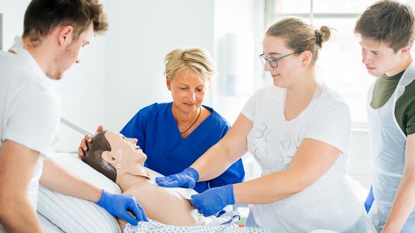 Eine Gruppe von fünf Personen übt an einem menschlichen Trainingssimulator im Krankenhaus. Zwei Personen fassen den Simulator an, während eine weitere Person Anweisungen gibt. Alle tragen Handschuhe und konzentrieren sich auf die medizinische Ausbildung.
