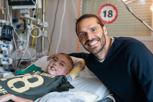 Ein lächelnder Mann sitzt neben einem kranken Jungen im Krankenhausbett. Der Junge trägt ein grünes T-Shirt mit der Zahl 18. Beide wirken glücklich und verbunden, was eine Botschaft von Hoffnung und Unterstützung vermittelt.