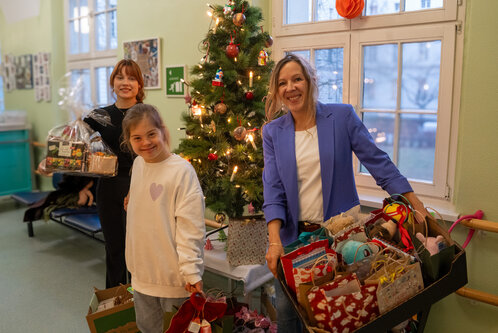Drei Frauen stehen in einem Raum mit einem beleuchteten Weihnachtsbaum. Eine Frau hält einen Geschenkkorb, während eine andere mit einem Korb voller Geschenke lächelt. Die dritte Frau, eine junge Person, steht neben ihnen und lächelt ebenfalls, alle wirken fröhlich und feiern die Weihnachtszeit.