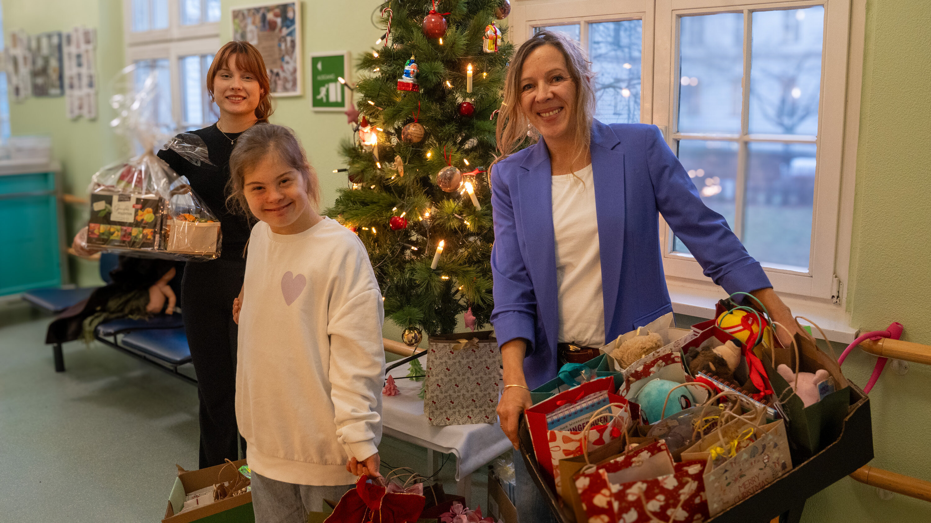 Drei Frauen stehen in einem Raum mit einem beleuchteten Weihnachtsbaum. Eine Frau hält einen Geschenkkorb, während eine andere mit einem Korb voller Geschenke lächelt. Die dritte Frau, eine junge Person, steht neben ihnen und lächelt ebenfalls, alle wirken fröhlich und feiern die Weihnachtszeit.