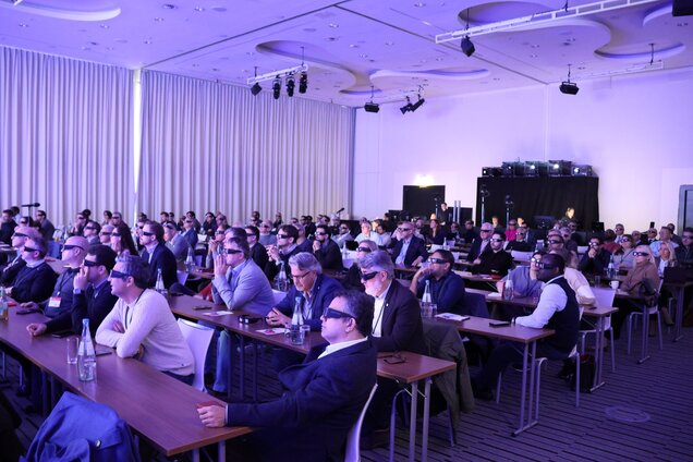 Eine große Gruppe von Menschen sitzt in einem Konferenzraum. Die Teilnehmer tragen Sonnenbrillen und blicken aufmerksam nach vorne. Tische und Wasserflaschen sind sichtbar, während eine Bühne im Hintergrund vorhanden ist. Die Atmosphäre ist ernst und konzentriert.