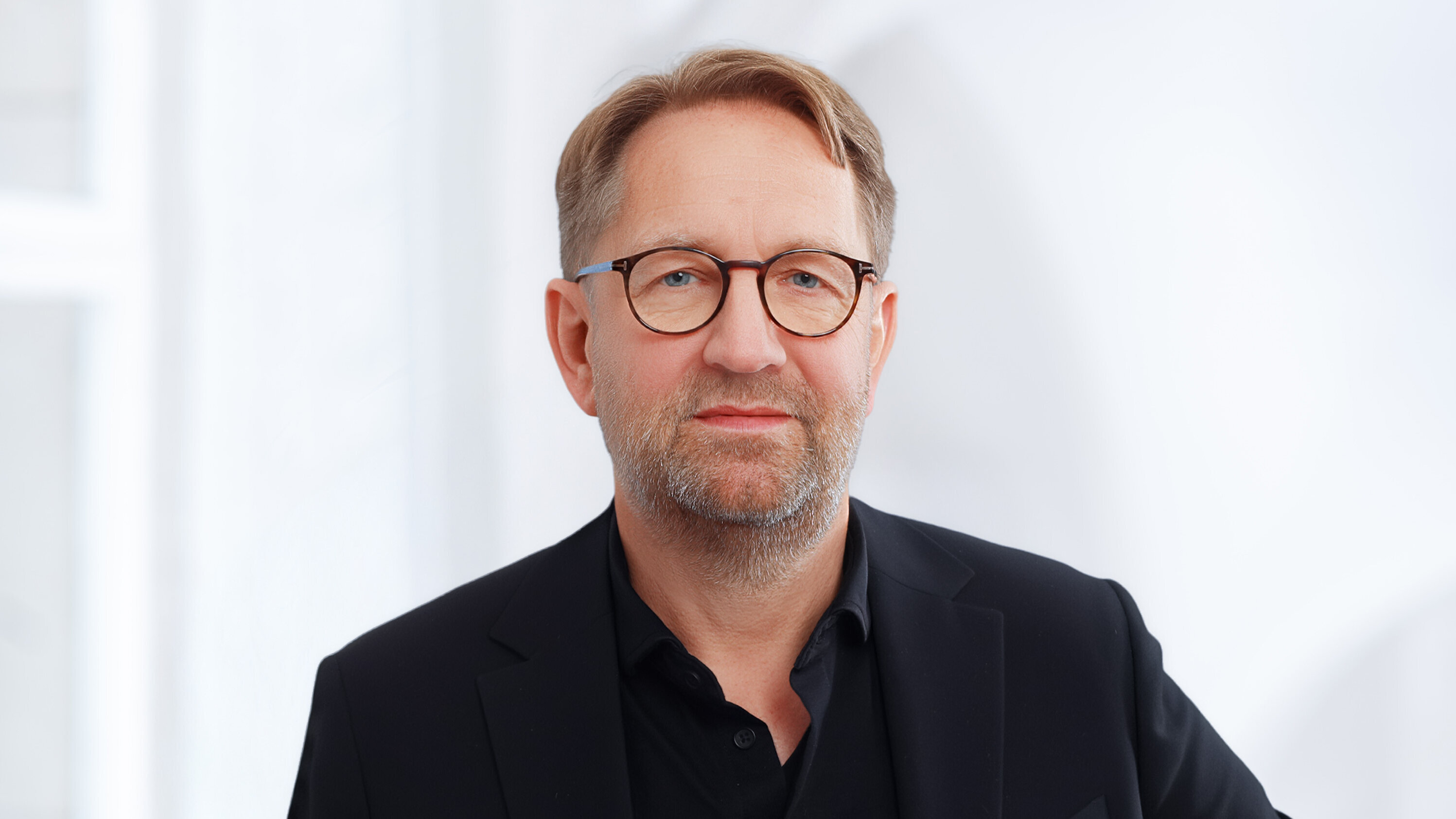 Ein Mann mit Brille und mittellangem, leicht grauem Haar trägt einen schwarzen Anzug und schaut direkt in die Kamera. Der Hintergrund ist hell und verschwommen, was den Fokus auf den Mann lenkt.