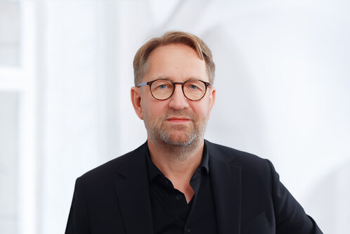 Ein Mann mit Brille und mittellangem, leicht grauem Haar trägt einen schwarzen Anzug und schaut direkt in die Kamera. Der Hintergrund ist hell und verschwommen, was den Fokus auf den Mann lenkt.