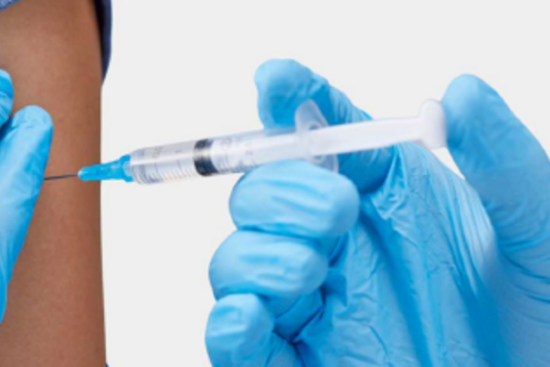 Eine Hand in blauen Handschuhen hält eine Spritze, die kurz davor ist, in den Oberarm einer anderen Person injiziert zu werden. Der Fokus liegt auf der Spritze und dem Arm, was den Impfprozess symbolisiert.