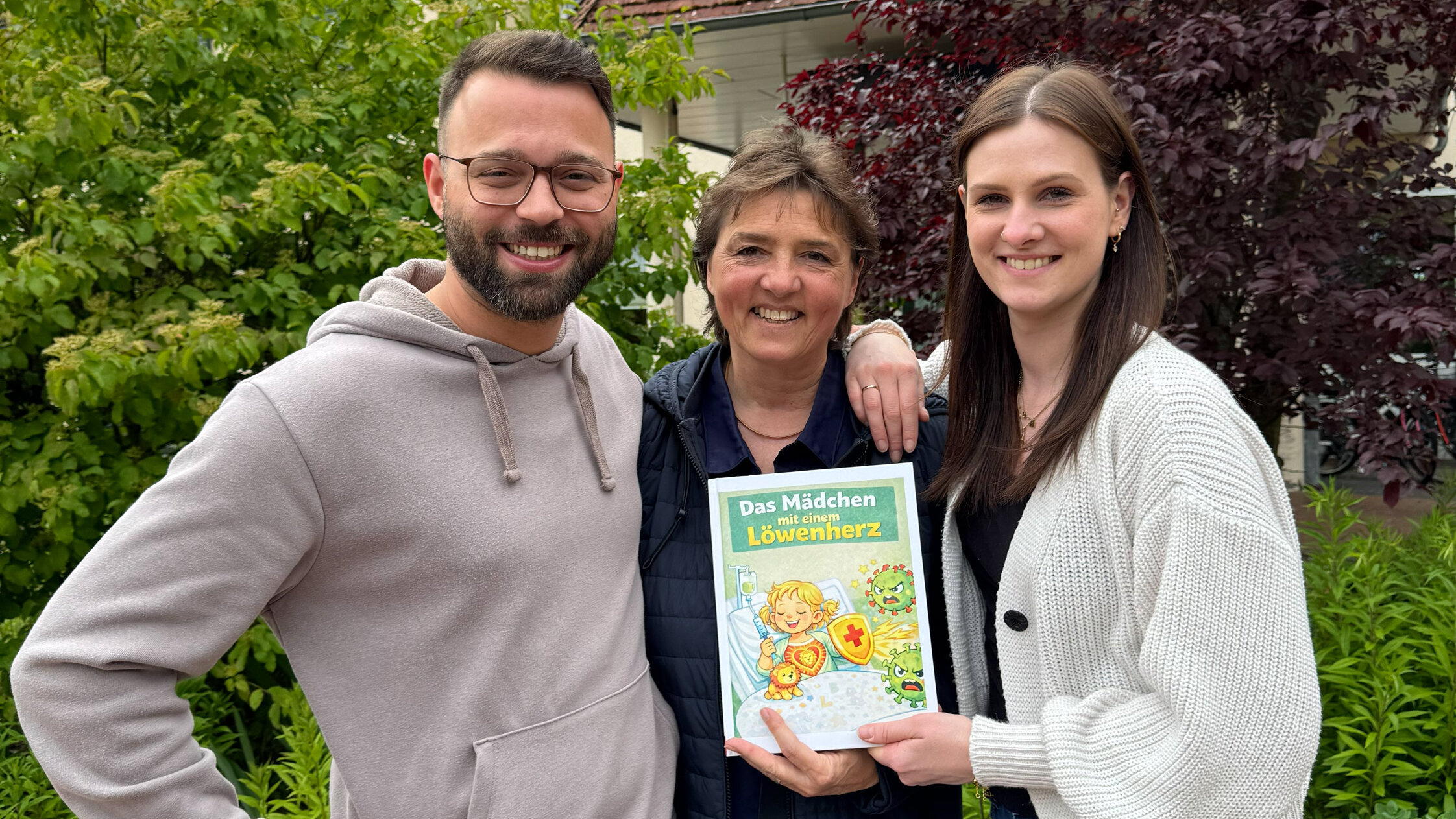 Drei Personen stehen im Freien, umgeben von grünen Pflanzen. Sie lächeln und halten ein Kinderbuch mit dem Titel „Das Mitteln von Löwenzahn“ in den Händen. Im Hintergrund sind Teile eines Hauses sichtbar.