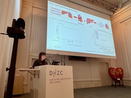 Eine Vortragende steht an einem Pult und präsentiert Folien über Herz- und Lebertransplantationen. Die Folien zeigen Diagramme und Informationen zu kombinierten Transplantationen und den Langzeitergebnissen. Im Hintergrund ist ein dekoratives Herz sichtbar. Der Vortrag findet im Deutschen Herzzentrum statt.