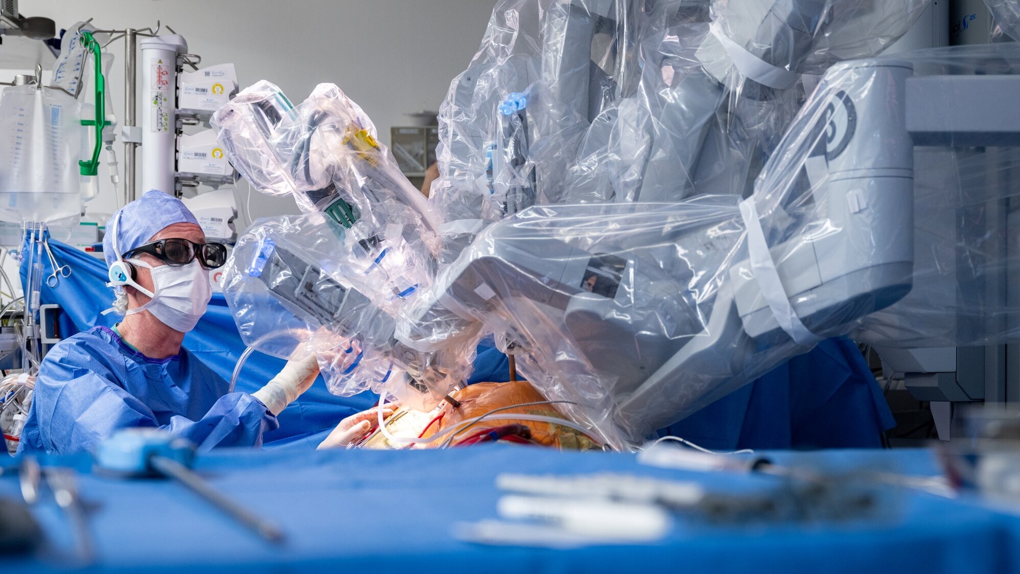 Ein Chirurg in Schutzkleidung führt eine Operation mit einem Roboter durch. Der Roboterarm ist durch eine transparente Hülle geschützt und über dem Patienten positioniert. Instrumente und medizinische Geräte sind im Hintergrund sichtbar, während der Fokus auf dem chirurgischen Eingriff liegt.