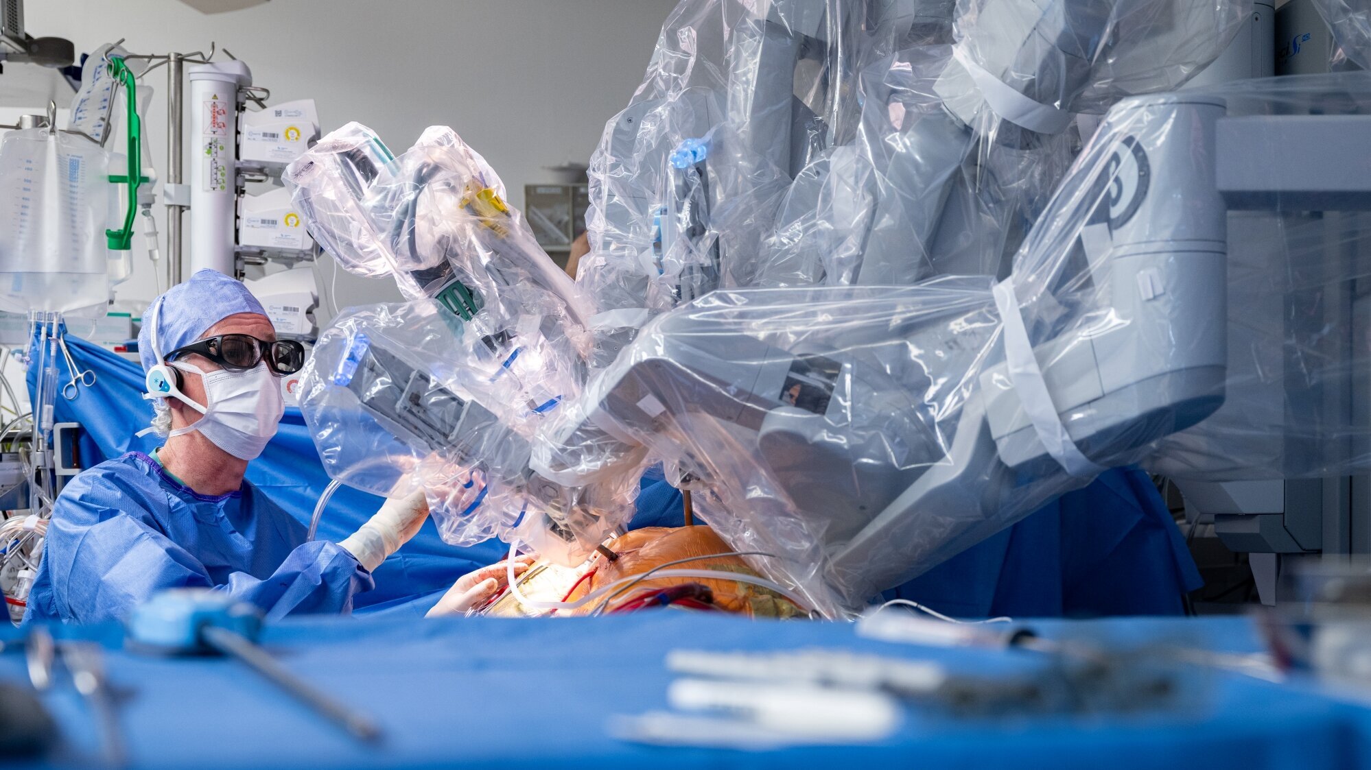 Ein Chirurg in Schutzkleidung führt eine Operation mit einem Roboter durch. Der Roboterarm ist durch eine transparente Hülle geschützt und über dem Patienten positioniert. Instrumente und medizinische Geräte sind im Hintergrund sichtbar, während der Fokus auf dem chirurgischen Eingriff liegt.