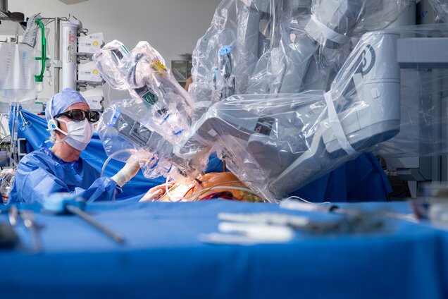 Ein Chirurg in Schutzkleidung führt eine Operation mit einem Roboter durch. Der Roboterarm ist durch eine transparente Hülle geschützt und über dem Patienten positioniert. Instrumente und medizinische Geräte sind im Hintergrund sichtbar, während der Fokus auf dem chirurgischen Eingriff liegt.