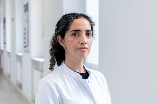 Eine Frau mit lockigem, dunklem Haar steht in einem hellen, modernen Flur. Sie trägt einen weißen Kittel. Ihr Blick ist ernst und aufmerksam. Im Hintergrund sind unscharfe Fenster und Wände sichtbar, die einen medizinischen Kontext vermitteln.
