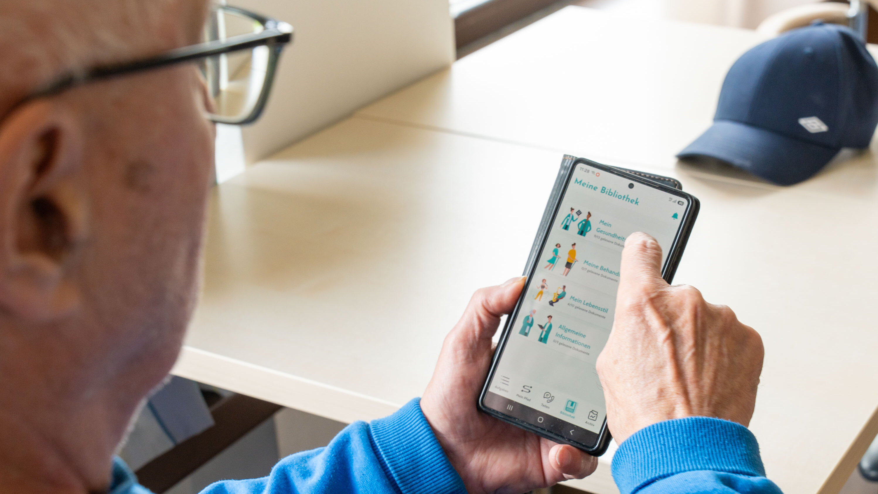 Ein älterer Mann mit Brille hält ein Smartphone in der Hand und schaut darauf. Er tippt mit dem Finger auf den Bildschirm, der eine App mit verschiedenen Icons und Informationen anzeigt. Im Hintergrund ist ein Tisch zu sehen.
