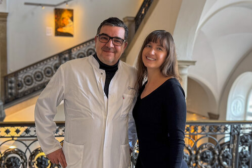 Zwei Personen stehen vor einem geschwungenen Geländer in einem eleganten Innenraum. Der Mann trägt einen weißen Laborkittel und Brille, die Frau trägt ein schwarzes Oberteil und lächelt. Im Hintergrund ist eine Treppe und ein Kunstwerk an der Wand sichtbar.