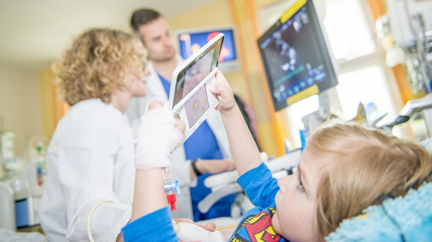 Ein Kind liegt im Krankenhausbett und hält ein Tablet in der Hand. Auf dem Bildschirm ist eine Anwendung zu sehen. Im Hintergrund sind zwei Ärzte zu sehen, die miteinander sprechen und auf einen Monitor mit medizinischen Daten schauen.
