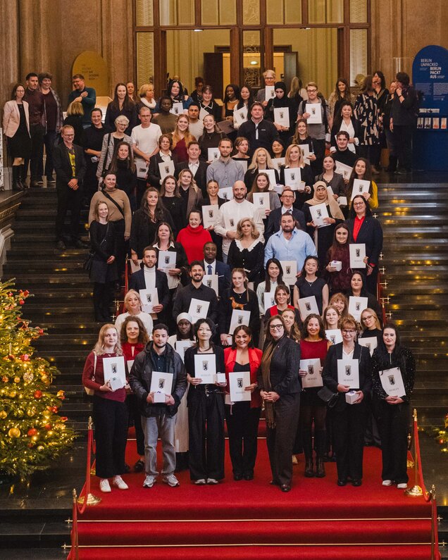 Eine große Gruppe von Menschen steht auf einer Treppe, die mit rotem Teppich ausgelegt ist. Sie halten Urkunden in den Händen und lächeln. Im Hintergrund sieht man eine festlich dekorierte Umgebung mit einem Weihnachtsbaum.