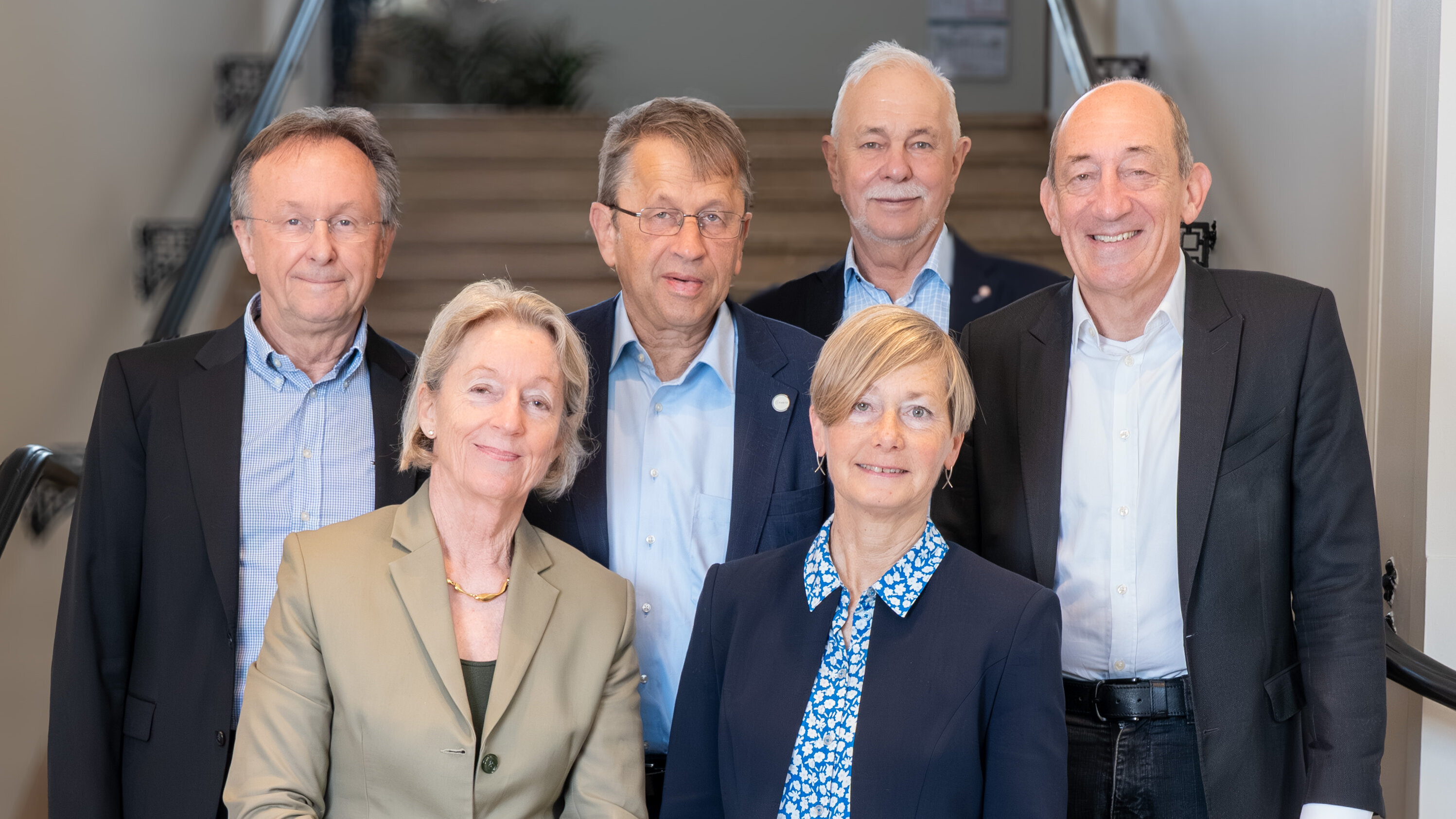 Eine Gruppe von sechs Personen steht auf einer Treppe. Sie sind professionell gekleidet und blickens in die Kamera. Im Hintergrund sind Stufen und eine Wand sichtbar. Die Personen scheinen miteinander zu kommunizieren oder in einer besprechenden Atmosphäre zu sein.