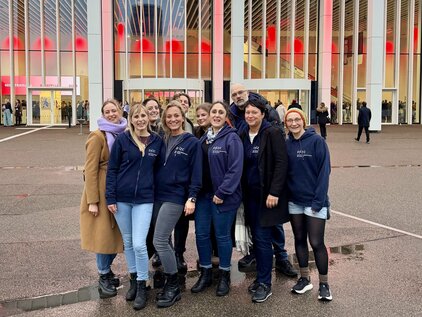 Eine Gruppe von neun Personen steht lächelnd vor einem großen Gebäude mit einem Banner, das "Herzlich Willkommen! HOTSPOT HERZMEDIZIN 2016" zeigt. Die Frauen tragen dunkelblaue Kleidung und bilden einen freundlichen Kreis. Im Hintergrund sind weitere Messebesucher und das Gebäude zu sehen.