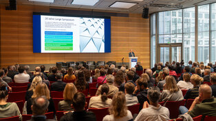 Ein Symposium mit dem Titel „20 Jahre Lange Symposium“ findet in einem modern ausgestatteten Saal statt. Ein Referent präsentiert Inhalte auf einer Leinwand, während ein Publikum aus Fachleuten ihm aufmerksam zuhört. Thema sind Entwicklungen in der Herzmedizin und digitale Transformation.